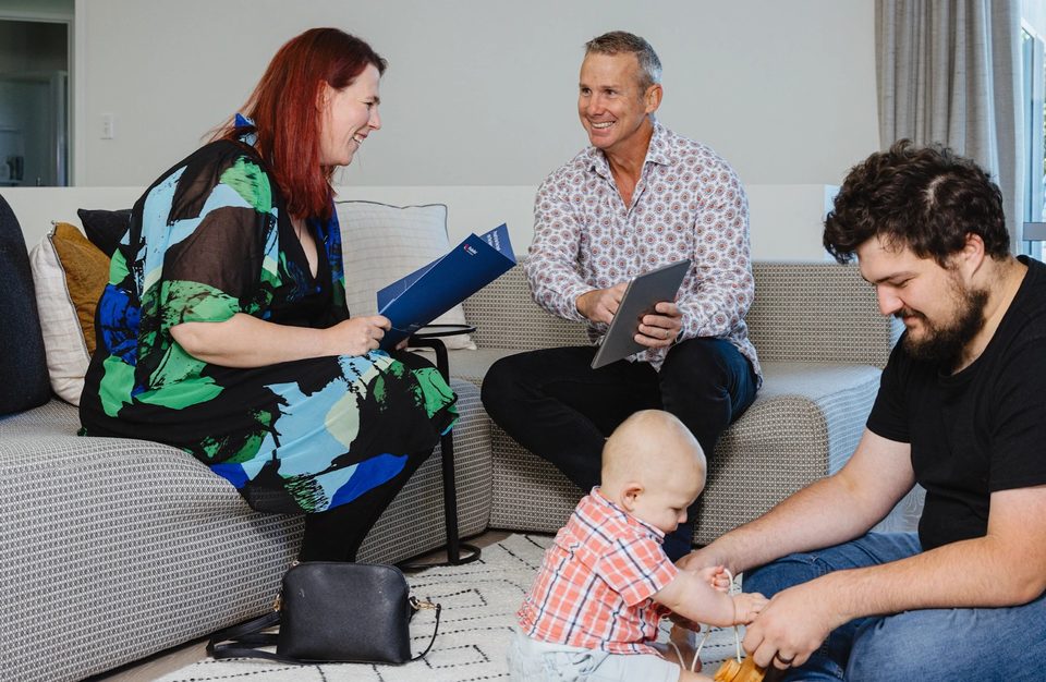 A Pukeko rental manager holding a blue folder talking with a couple and their baby in a lounge.