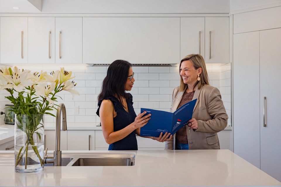 Two women reviewing paperwork in a blue Pukeko management folder at a kitchen bench.