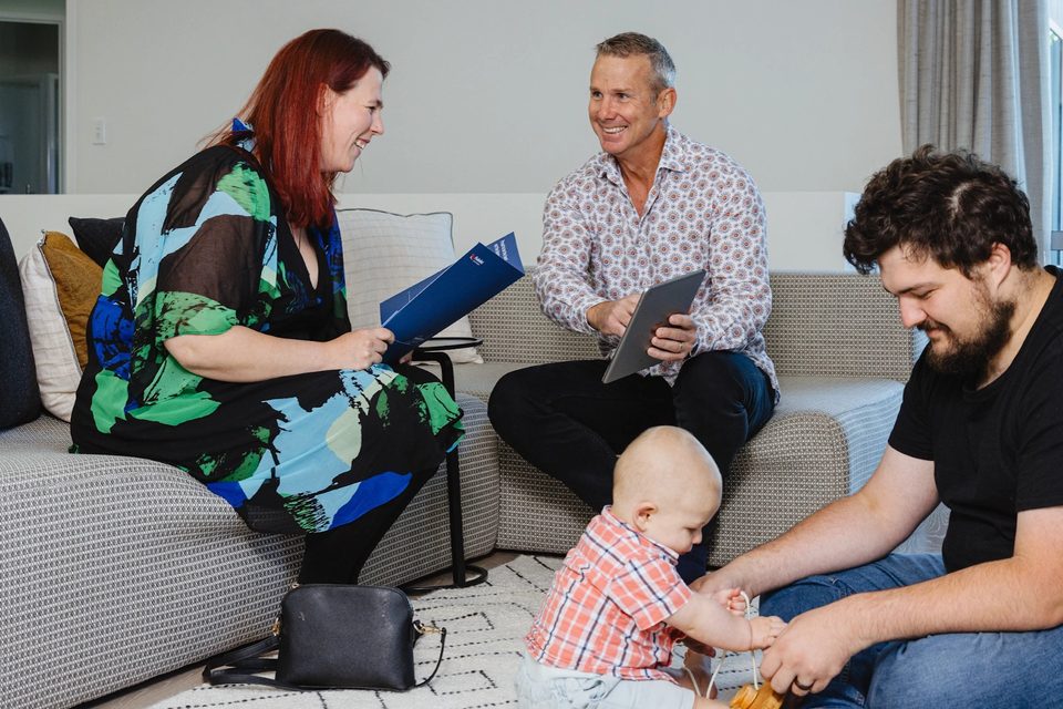 A rental manager with a blue folder sitting with a couple and their baby in a lounge.